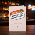 A novelty greeting card standing upright on a wooden table, with a blurred bar background. The card has a light pink background and a speech-bubble style label that reads 'Hi! I'm.