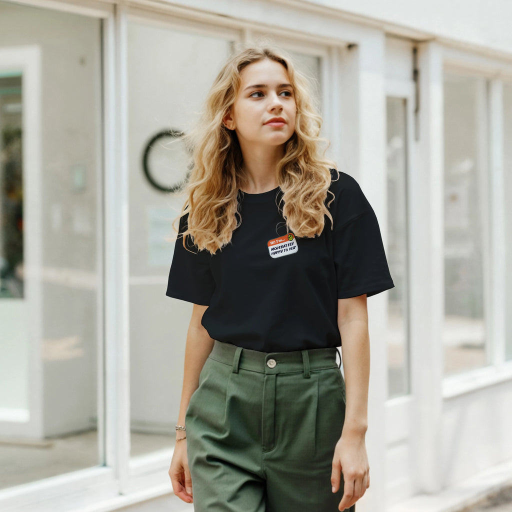 Lifestyle image of a young woman wearing a black t-shirt with a printed imitation nametag saying 'Hi! I'm Moderately Here to Help' by Retailer Therapy