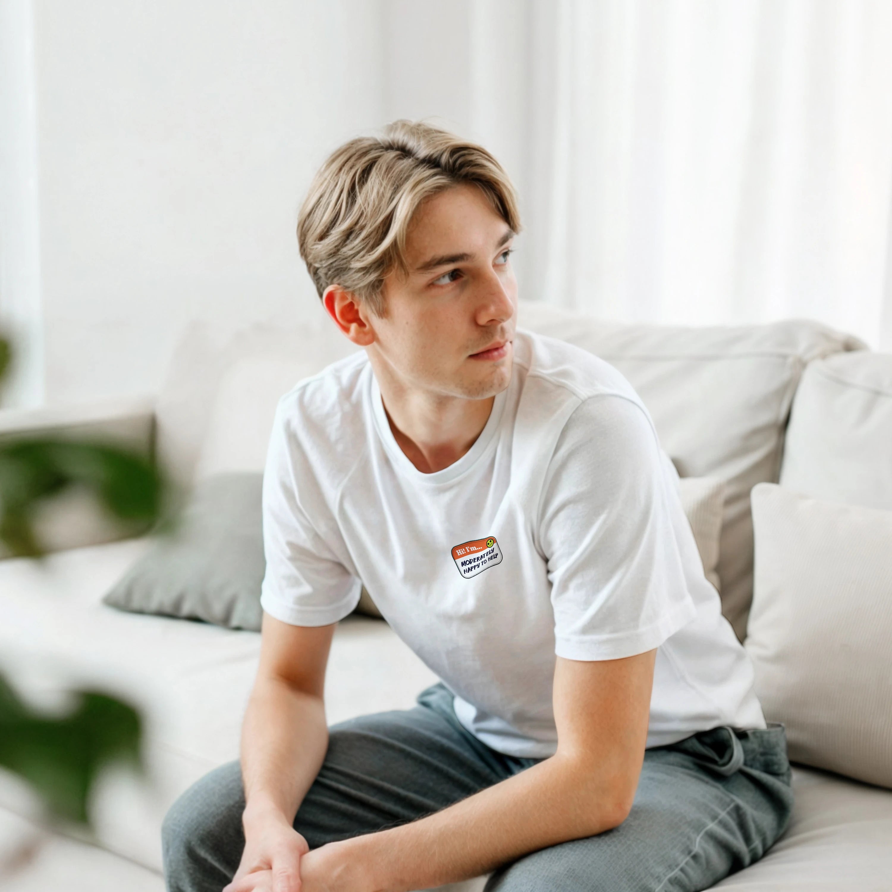 Lifestyle image of a young man wearing a black t-shirt with a printed imitation nametag saying 'Hi! I'm Moderately Here to Help' by Retailer Therapy