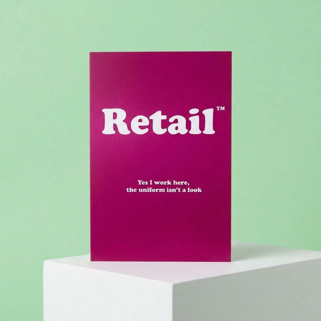 A magenta greeting card with white text reading 'Retail' and 'Yes I work here, the uniform isn't a look', displayed on a white pedestal against a mint green background.