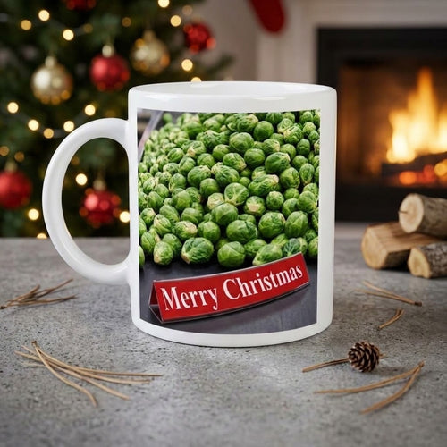 A white ceramic mug with a printed image of green Brussels sprouts and a red banner reading 'Merry Christmas', set on a gray stone surface with a blurred Christmas tree and fireplace in the background.