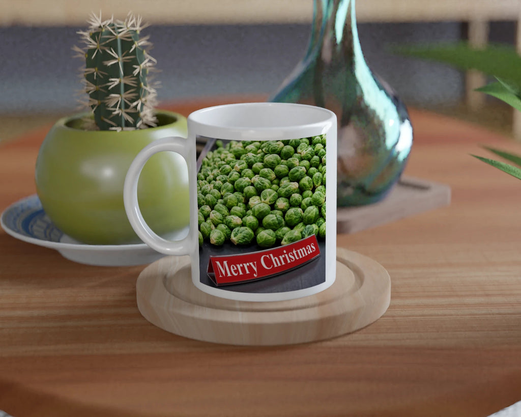 A white ceramic mug with a printed image of green peas and a red banner reading 'Merry Christmas' sits on a round wooden coaster on a wooden table. A small potted cactus and a blue glass vase are in the background.